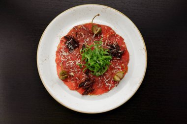 Dana fileto carpaccio, roka ve kapari, bir tabakta, koyu bir arka planda.