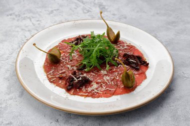 Dana fileto carpaccio, roka ve kapari, tabakta, hafif arkaplanda.