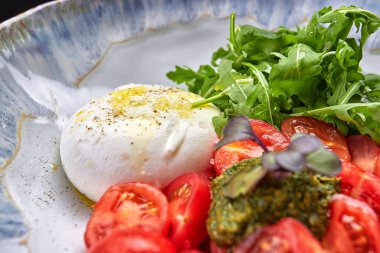 Roka, vişneli domates ve pesto soslu Burrata. Işıklı bir plakada, siyah bir arkaplanda.