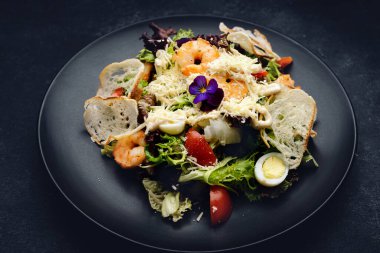 Karides ve çiçekli Sezar salatası, siyah bir tabakta, koyu betonda.