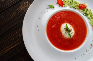 Tahta arka planda beyaz bir tabakta domates çorbası, domates ve otlarla.