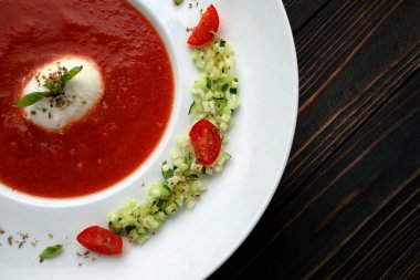 Tahta arka planda beyaz bir tabakta domates çorbası, domates ve otlarla.