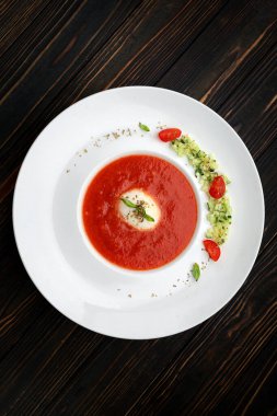 Tahta arka planda beyaz bir tabakta domates çorbası, domates ve otlarla.