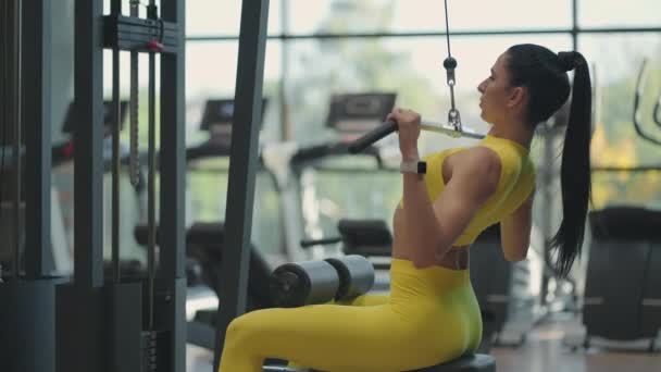 Een jonge brunette in een geel pak doet oefeningen voor de spieren van de rug op de simulator. Een Spaanse vrouw trekt een touw aan de simulator. Gesimuleerde pull-ups — Stockvideo