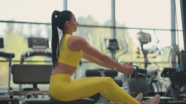 Spaanse vrouw doet oefeningen op de roeimachine. Een meisje zit achter een simulator en heet een roeimachine. zitten trekt het gewicht van grote ramen en loopbanden van de sportschool — Stockvideo