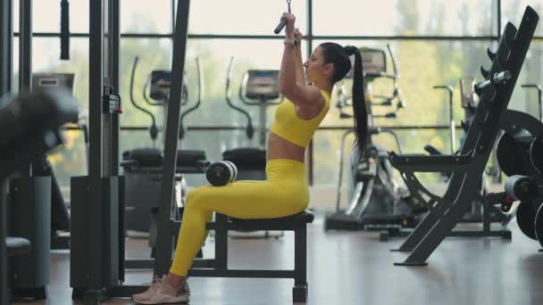 一位穿着黄色西服的年轻的黑发女子正在模拟器上锻炼背部肌肉。西班牙女人把绳子拉到模拟器上.模拟拉力 — 图库视频影像