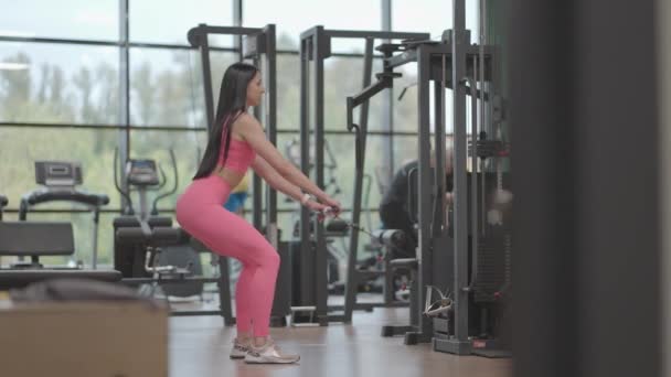 A young woman squats in a crossover to train her thigh and leg muscles. Buttocks training. Professional female athlete does leg exercises squats — Stock Video