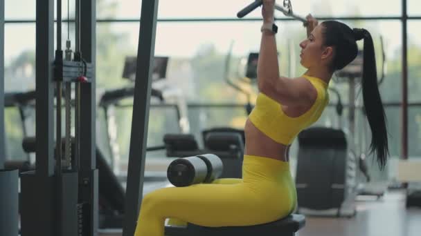 스페인 여자가 헬스클럽 에서 시뮬레이터 에 앉아 몸무게때문에 금속 로프 를 등 의근 육을들어올리고 있다. 갈색 머리 여자가 시뮬레이터를 작동 합니다. 등 근육 시뮬레이터 운동을 하는 모습 — 비디오