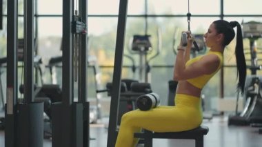 Spor salonunda simülatörde oturan İspanyol kadın sırtında ağırlık pompalarıyla metal bir ip çekiyor. Esmer kadın simülatörü çekiyor. Sırt kası simülatörü için egzersiz yapıyor
