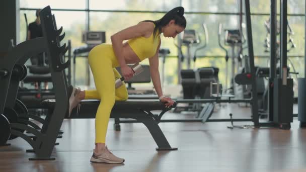 Іспанська спортсменка у спортивній манері тренується з німим дзвоном в одній руці, спираючись на лавку в фітнес-залі — стокове відео