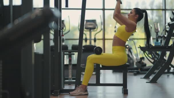 Hispanic woman sitting on a simulator in the gym pulls a metal rope with the weight pumps up the muscles of the back. brunette woman pulls on simulator. performing exercise for back muscles simulator — Stock Video