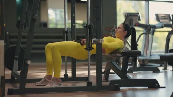 Una joven con un chándal amarillo levanta su cuerpo con una barra para entrenar la parte posterior del muslo y las nalgas. Entrenamiento de pesas en el simulador de herrero. Levantar la cintura y el cuerpo con peso en el gimnasio — Vídeos de Stock