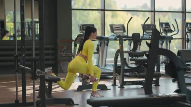 Mujer deportiva está haciendo saltos para las piernas con mancuernas en las manos en el gimnasio. Joven mujer hispana musculosa haciendo ejercicios con pesas en el gimnasio en ropa deportiva amarilla — Vídeos de Stock