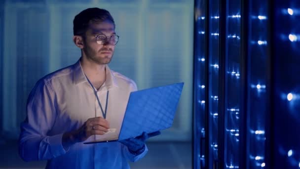 Hombre ingeniero de red haciendo una comprobación del sistema de pie en la sala de servidores con su computadora portátil. En el centro de datos, los especialistas en servidores de los hombres inspeccionan el sistema de trabajo y el hardware de los gabinetes informáticos del servidor rack — Vídeo de stock