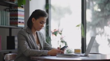 Tarz sahibi genç bir kadın internette sörf yaparak modern kafedeki bir masada oturuyor ve gülümsüyor. Ofisteki genç iş kadını cep telefonu kullanıyor.