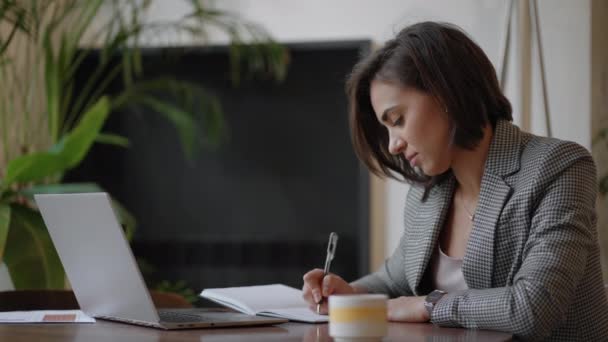 İş kadını grafiği analiz ediyor ve dizüstü bilgisayarda daktilo yazıyor. Uzak iş, gözlüklü genç bir kadın not defterine ve klavyeye not yazıyor. — Stok video