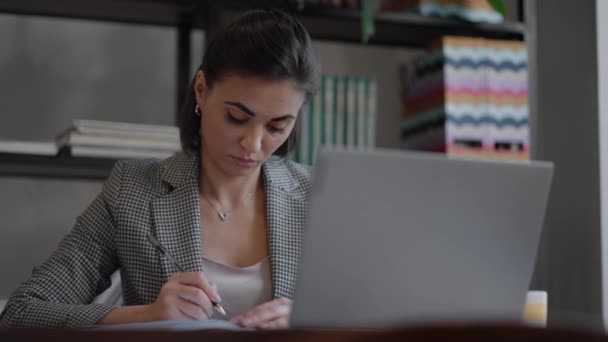 Επιχειρηματίας αναλύοντας το γράφημα και πληκτρολογώντας στο laptop. Απομακρυσμένη δουλειά, νεαρή γυναίκα με γυαλιά γράφει σημειώσεις στο πρόχειρο και πληκτρολογεί στο πληκτρολόγιο laptop — Αρχείο Βίντεο