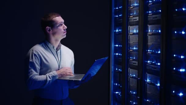 Young man holds device in hand and looks at screen, inspects equipment or hardware rack. Male programmer working with laptop and supporting service while standing in data center spbas. — Stock Video