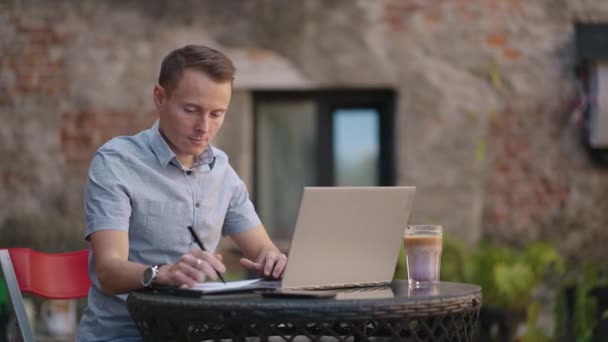 Un hombre sentado en un café de la calle trabaja remotamente escribiendo un bolígrafo en un cuaderno. Trabaja en el portátil. Un hombre trabaja remotamente en un portátil — Vídeo de stock