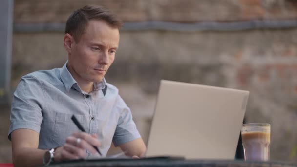 Een man die in een café zit, schrijft op afstand een pen in een notitieboekje. Werk op de laptop. Een man werkt op afstand op een laptop. — Stockvideo