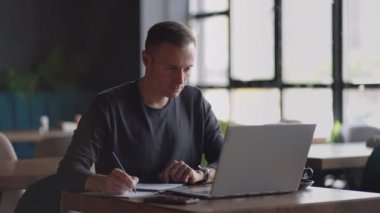 Mutfakta dizüstü bilgisayar kullanarak çalışan gülümseyen bir adam. Uzaktan çalışma ve uzaktan öğrenme. Karantinada kendini izole ederken uzaktan çalışma, adam finansal belgelerle çalışıyor.