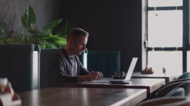 Mutfakta dizüstü bilgisayar kullanarak çalışan gülümseyen bir adam. Uzaktan çalışma ve uzaktan öğrenme. Karantinada kendini izole ederken uzaktan çalışma, adam finansal belgelerle çalışıyor.