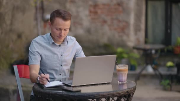 Muž sedící v pouliční kavárně pracuje na dálku a píše pero do sešitu. Pracuj v laptopu. Muž pracuje na dálku na notebooku — Stock video