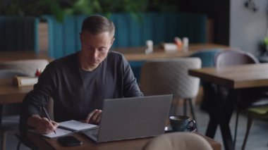 Mutfakta dizüstü bilgisayar kullanarak çalışan gülümseyen bir adam. Uzaktan çalışma ve uzaktan öğrenme. Karantinada kendini izole ederken uzaktan çalışma, adam finansal belgelerle çalışıyor.