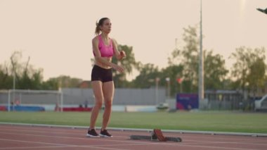 Düşüncelere dalmış genç bir kadın sporcu ısınıyor ve güneşli bir günde stadyumun koşu bandında koşuya hazırlanıyor.