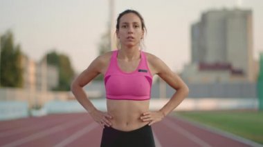 Düşüncelere dalmış genç bir kadın sporcu ısınıyor ve güneşli bir günde stadyumun koşu bandında koşuya hazırlanıyor.