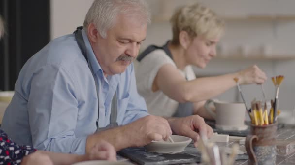 Μια ομάδα ηλικιωμένων σε ένα master class κεραμικής μαζί σμιλεύουν και κόβουν ένα σχέδιο σε κύπελλα από πηλό για την κατασκευή κεραμικών πιάτων — Αρχείο Βίντεο
