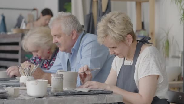 A group of elderly people at a master class in pottery together sculpt and cut a drawing on cups of clay for the manufacture of ceramic dishes — Stock Video