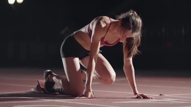 スタジアムの暗闇の中で一人の女性がコース上でレースを始める準備をしている。スローモーションの女性は暗闇のスタジアムで飢えのストライキから始まります — ストック動画