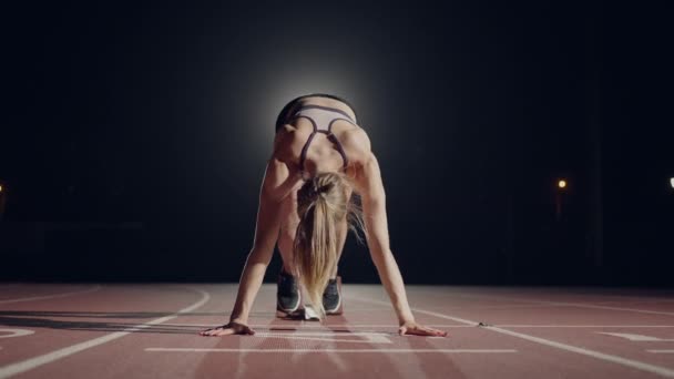 Una mujer en la oscuridad en el estadio se está preparando para comenzar una carrera en la pista. Mujer de cámara lenta comienza con huelgas de hambre en el estadio en la oscuridad — Vídeo de stock