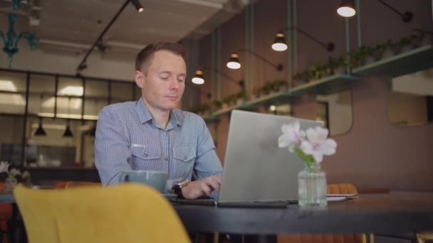 Gömlekli genç bir adam elinde dizüstü bilgisayarla bir masada oturuyor ve klavyeye yazı yazıyor. Bir öğrenci uzaktan çalışabilir. Bir iş adamı işini uzaktan yürütür. — Stok video