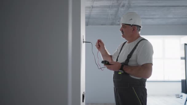 男性電気技師は、建設現場にスマートハウスを設置する準備をしているワイヤーテスターでネットワーク内の電圧をチェックします。. — ストック動画