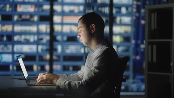 Trabalho noturno um homem se senta em uma mesa e imprime em um laptop contra o fundo da cidade noturna — Vídeo de Stock