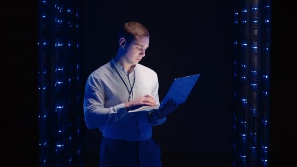 Ειδικός πληροφορικής εργάζεται με ένα φορητό υπολογιστή στην αίθουσα του διακομιστή κοιτάζοντας τα ντουλάπια με διακομιστές — Αρχείο Βίντεο