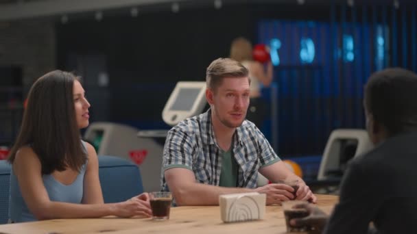Eine multiethnische Gruppe von Freunden sitzt gemeinsam an einem Tisch in einer Bowlingbahn und spricht — Stockvideo