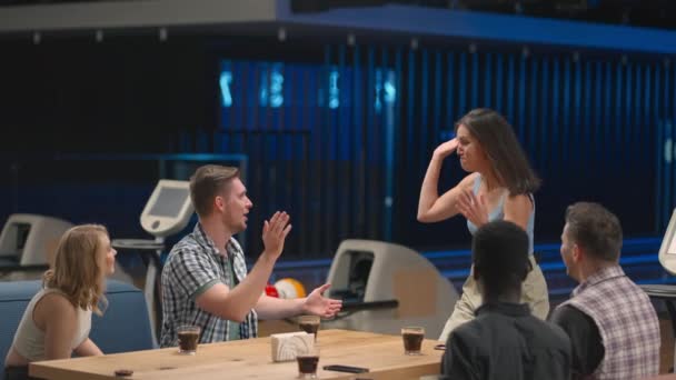 Eine fröhliche brünette Frau läuft auf Freunde zu und gibt fünf Männern und Frauen, die an einem Tisch in einem Bowlingclub sitzen. Feiern Sie Sieg und Siegesserie. Um zu schneiden und zum Sieg zu gratulieren. Teamerfolg. — Stockvideo