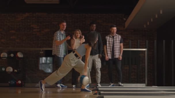 Una joven morena hace el lanzamiento final con una bola de bolos y gana el juego con el apoyo y la alegría de sus amigos de diferentes nacionalidades. Alégrate y celebra la victoria — Vídeo de stock