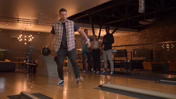 Een man in een bowlingbaan gooit een bal op de baan en slaat een schot in slow motion en springt en danst van vreugde. Vrienden fans. Een groep multi-etnische vrienden spelen samen bowlen. — Stockvideo