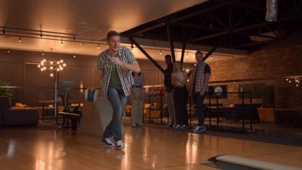 A man in a bowling alley throws a ball on the track and knocks out a shot in slow motion and jumps and dances for joy. A group of multi-ethnic friends play bowling together clapping happy, supporting — Stock Video