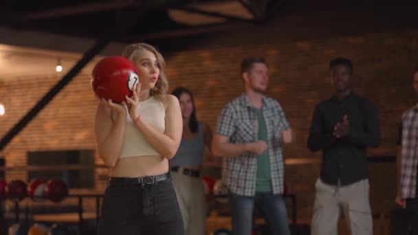 Porträt Eine junge blonde Frau macht den letzten Wurf mit einer Bowlingkugel und gewinnt das Spiel mit der Unterstützung und Freude ihrer Freunde verschiedener Nationalitäten. Jubeln und den Sieg feiern — Stockvideo