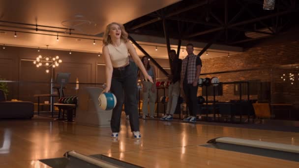 Uma jovem no bowling joga uma bola na pista e cai no chão em câmera lenta e salta e dança de alegria. Amigos fãs apoiam e aplaudem. Amigos multi-étnicos jogam bowling juntos. — Vídeo de Stock