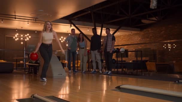 A young woman in bowling throws a ball on the track and knocks out a shot in slow motion and jumps, dances for joy. Friends fans support, clap. A group of multi-ethnic friends play bowling together. — Stock Video