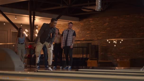 En svart afroamerikansk man kastar en bowlingboll och slår ut en bild med ett kast och kramar och gläds med vänner. Multietnisk grupp vänner bowling — Stockvideo