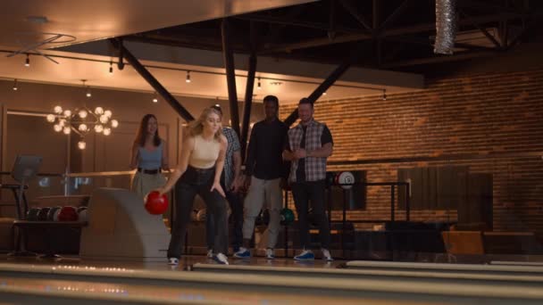 Una bella bionda lancia una palla da bowling e tira fuori un tiro con un solo tiro e abbraccia e gioisce con i suoi amici. Gruppo multietnico di amici bowling — Video Stock