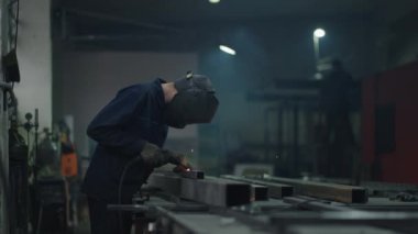 Meşaleli Endüstriyel Kaynakçı. Kaynak maskesi ve kaynakçı derileri, inşaat alanında kaynak makinesi ile kaynak yapan metaller, kenarlarda mavi kıvılcımlar uçuşuyor.