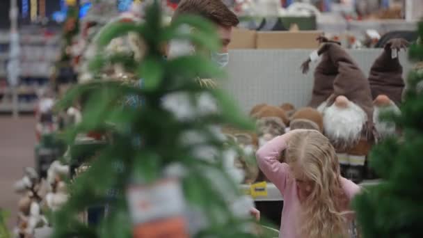 Pai e filha caminham para o supermercado com uma máscara protetora, a menina monta em um carrinho de compras, escolhendo uma árvore de Natal — Vídeo de Stock